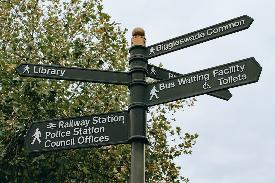 A black multi-directional street signpost situated outdoors during daytime, mounted on a metal pole with a rounded top, surrounded by green trees with some leaves beginning to change colour. The signpost features several directional signs with white text and icons, including signs pointing to Biggleswade Common, Library, Railway Station, Police Station, Council Offices, Bus Waiting Facility, and Toilets. The signs are attached to horizontal arms extending from the central pole, which is mounted on a concrete base. The background sky is cloudy, providing diffused lighting. This street signage may be encountered during a home relocation or moving process, as part of understanding local infrastructure or planning logistics involved in house removals, potentially supported by Man with Van Oakleigh Park, a company specializing in removals and furniture transport in the area.