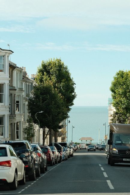 A residential street scene during daytime showing a row of parked cars along the curb, including sedans and SUVs in various colours, with large leafy trees lining both sides of the road. In the background, there is a view of a distant body of water, likely the coast, with a small pavilion or gazebo structure near the shoreline. A black van and other vehicles are positioned to the right, possibly involved in home relocation or furniture transport activities. The scene is well-lit with natural daylight, under a partly cloudy sky, providing an accurate depiction of an urban environment suitable for a house removal and moving service context, as handled by Man with Van Oakleigh Park.