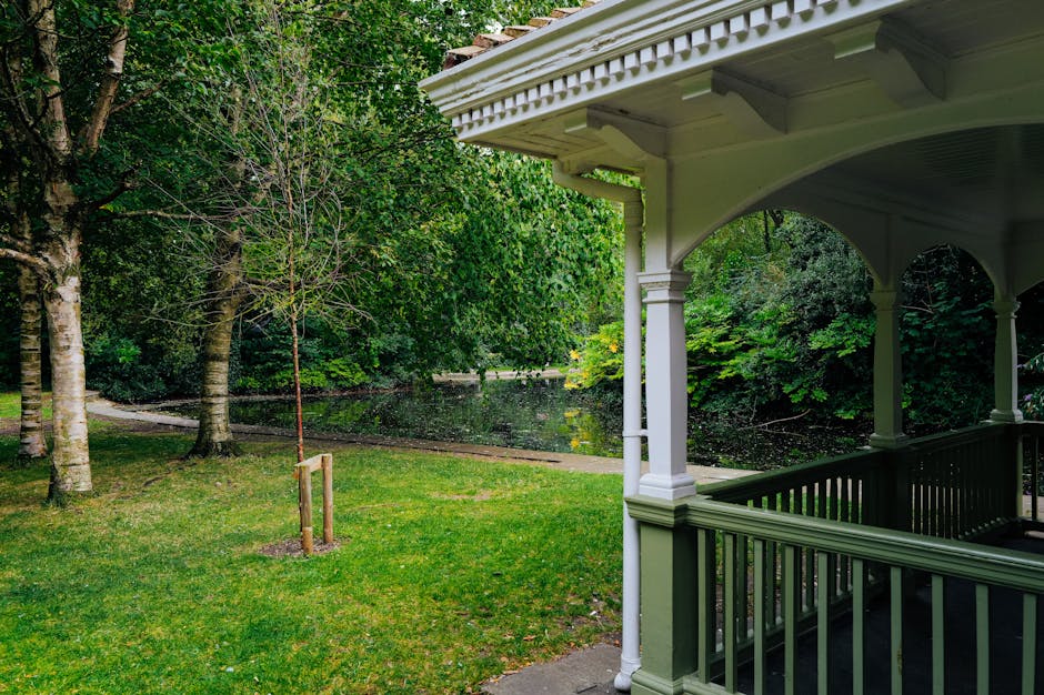 View from a residential porch overlooking a lush green garden with a small tree protected by a wooden stake, and a pond surrounded by trees and bushes. The porch has white painted wooden columns and a decorative railing, with the roof extending outward. Inside the garden, several mature trees with thick trunks and leafy canopies provide shade and greenery. The grass is well-maintained, and the scene suggests a peaceful outdoor environment suitable for home relocation preparations, with no visible moving boxes or furniture, indicating an outdoor setting possibly during the packing or transport process managed by Man with Van Oakleigh Park as part of their removals services.
