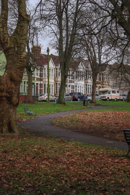 Image of a residential street with Victorian-style terraced houses in Oakleigh Park during daylight, viewed from a park with a paved pathway winding through grass and fallen leaves. Several parked cars, including a silver hatchback, dark sedan, and white van, are positioned along the street in front of the houses. Mature trees with bare branches line the sidewalk, suggesting late autumn or early winter. In the foreground, partially visible benches and a small black trash bin are situated on the grass beside the pathway. The scene captures a quiet suburban environment appropriate for home relocations and moving services, with no activity visible related to furniture transport or packing. The setting provides a clear view of the surroundings where Man with Van Oakleigh Park might facilitate house removals, including navigating tight lanes and outdoor loading areas.