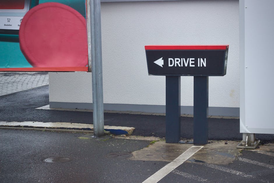 A rectangular black and red directional 'Drive In' sign with an arrow pointing left, mounted on two vertical black posts, positioned on a paved driveway outside a white building. To the left, part of a red circular sign and a metal pole are visible, along with a partial view of a brick wall and pavement. The area shows typical outdoor lighting with natural daylight. This scene relates to house removals and moving logistics, illustrating access points used during furniture transport and home relocation processes, with the sign indicating the direction for vehicles or delivery access. Man with Van Oakleigh Park's professional removals services often involve navigating such drive-in points for efficient loading and unloading of furniture and packed boxes in tight lanes and residential areas.