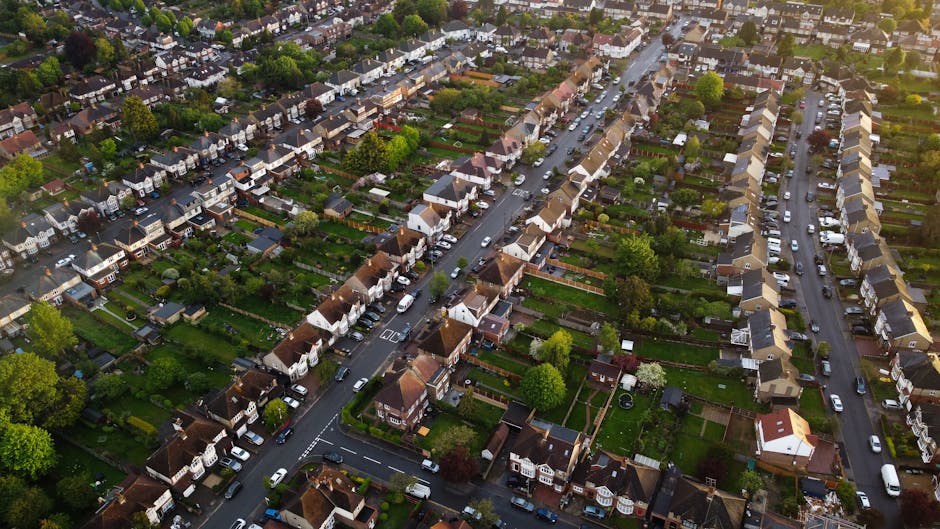 Image of a residential street with Victorian-style terraced houses in Oakleigh Park during daylight, viewed from a park with a paved pathway winding through grass and fallen leaves. Several parked cars, including a silver hatchback, dark sedan, and white van, are positioned along the street in front of the houses. Mature trees with bare branches line the sidewalk, suggesting late autumn or early winter. In the foreground, partially visible benches and a small black trash bin are situated on the grass beside the pathway. The scene captures a quiet suburban environment appropriate for home relocations and moving services, with no activity visible related to furniture transport or packing. The setting provides a clear view of the surroundings where Man with Van Oakleigh Park might facilitate house removals, including navigating tight lanes and outdoor loading areas.