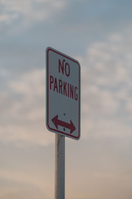 A vertical rectangular white 'No Parking' sign with red and black text, mounted on a metal pole. The sign features a double-headed red arrow at the bottom pointing left and right. It is positioned outdoors against a background of a cloudy sky during daytime, with some diffuse sunlight illuminating the scene. The sign is likely located on a street or parking area near a residential property, which is relevant to house removals and moving services offered by Man with Van Oakleigh Park. Its purpose is to indicate restrictions on parking in the vicinity, aligning with local regulations such as those under Barnet Council for Oakleigh Park. The image emphasizes outdoor traffic management signage relevant to relocating homes or transporting furniture and boxes within urban environments, supporting the context of home relocation and furniture transport logistics.