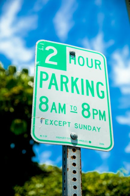 A green and white parking restriction sign mounted on a metal post beneath a bright blue sky with scattered clouds. The sign indicates a two-hour parking limit from 8 am to 8 pm, Monday to Saturday, with an exception for Sundays. In the background, blurred green foliage is visible, suggesting an outdoor urban or suburban setting. The sign is part of local traffic regulation, relevant to household relocations and parking management during home removal services by Man with Van Oakleigh Park, supporting efficient planning of furniture transport and moving logistics in the Oakleigh Park area.