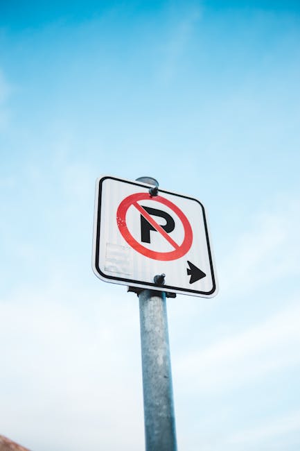 A vertical rectangular white 'No Parking' sign with red and black text, mounted on a metal pole. The sign features a double-headed red arrow at the bottom pointing left and right. It is positioned outdoors against a background of a cloudy sky during daytime, with some diffuse sunlight illuminating the scene. The sign is likely located on a street or parking area near a residential property, which is relevant to house removals and moving services offered by Man with Van Oakleigh Park. Its purpose is to indicate restrictions on parking in the vicinity, aligning with local regulations such as those under Barnet Council for Oakleigh Park. The image emphasizes outdoor traffic management signage relevant to relocating homes or transporting furniture and boxes within urban environments, supporting the context of home relocation and furniture transport logistics.