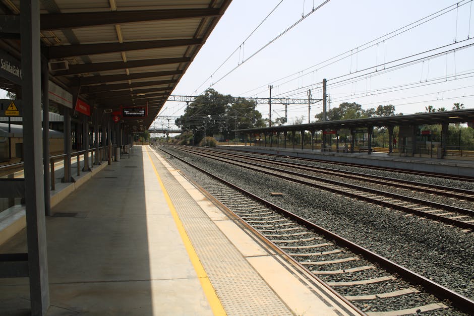 The image shows an empty train station platform with a covered waiting area on the left side, featuring a row of metal benches and a yellow safety line along the edge. The platform is illuminated by natural daylight, casting shadows from the roof structure onto the paved surface. Multiple railway tracks run parallel to the platform, with gravel beds supporting the rails. In the background, there are overhead electric wires and support structures, along with some distant trees and a partly cloudy sky. The environment appears quiet, with no trains or people present. This setting is related to transportation and logistics, supporting services such as house removals and furniture transport where efficient access and unloading are essential, which Man with Van Oakleigh Park provides as part of their house moving solutions.