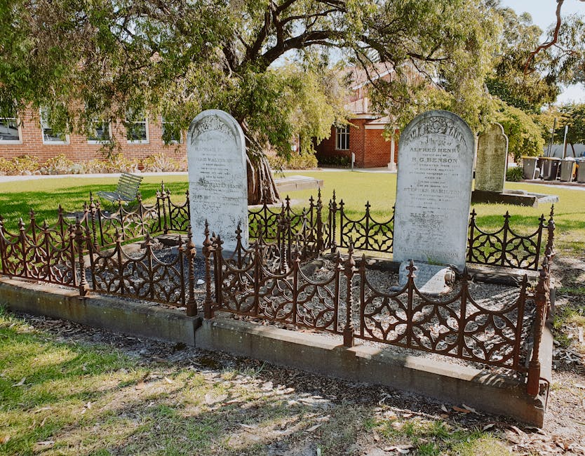 A small fenced grave site featuring two upright headstones made of light-colored stone, situated on a concrete platform within a landscaped area with green grass and trees. The grave is surrounded by an ornate, dark metal wrought iron fence with finials, which encloses the headstones and a section of gravel surface. The area appears to be part of a memorial garden or cemetery, with a park bench and a red-brick building visible in the background under a partly cloudy sky. The scene is well-lit, showing details of the fencing, headstones, and surrounding environment, and it reflects a typical setting for house removals and relocation services, such as loading or access planning, where clear and accurate visual descriptions are essential.