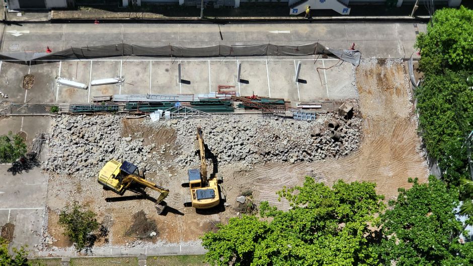 An aerial view of a construction site on a paved street in Oakleigh Park, showing a section of road closed off for repair. The area is cordoned with black safety barriers, and there are multiple const